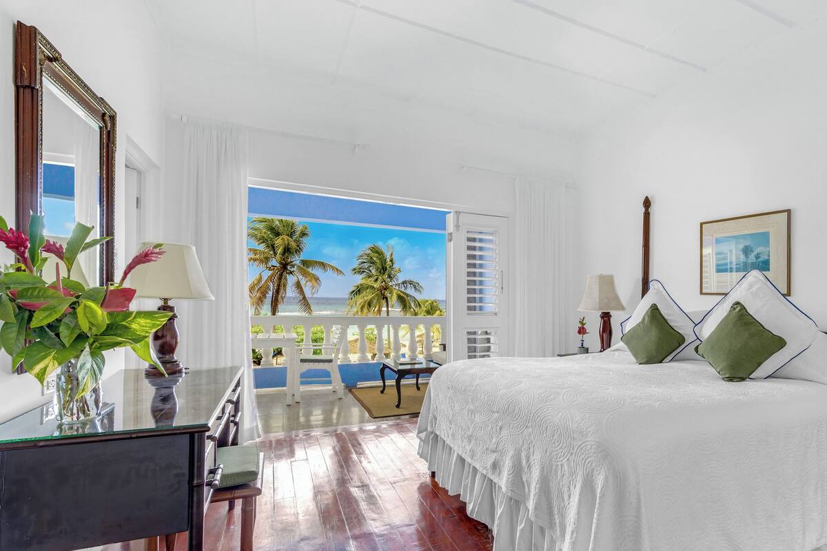 A hotel room with a large bed, a desk, a chair, and a view of palm trees and the ocean.