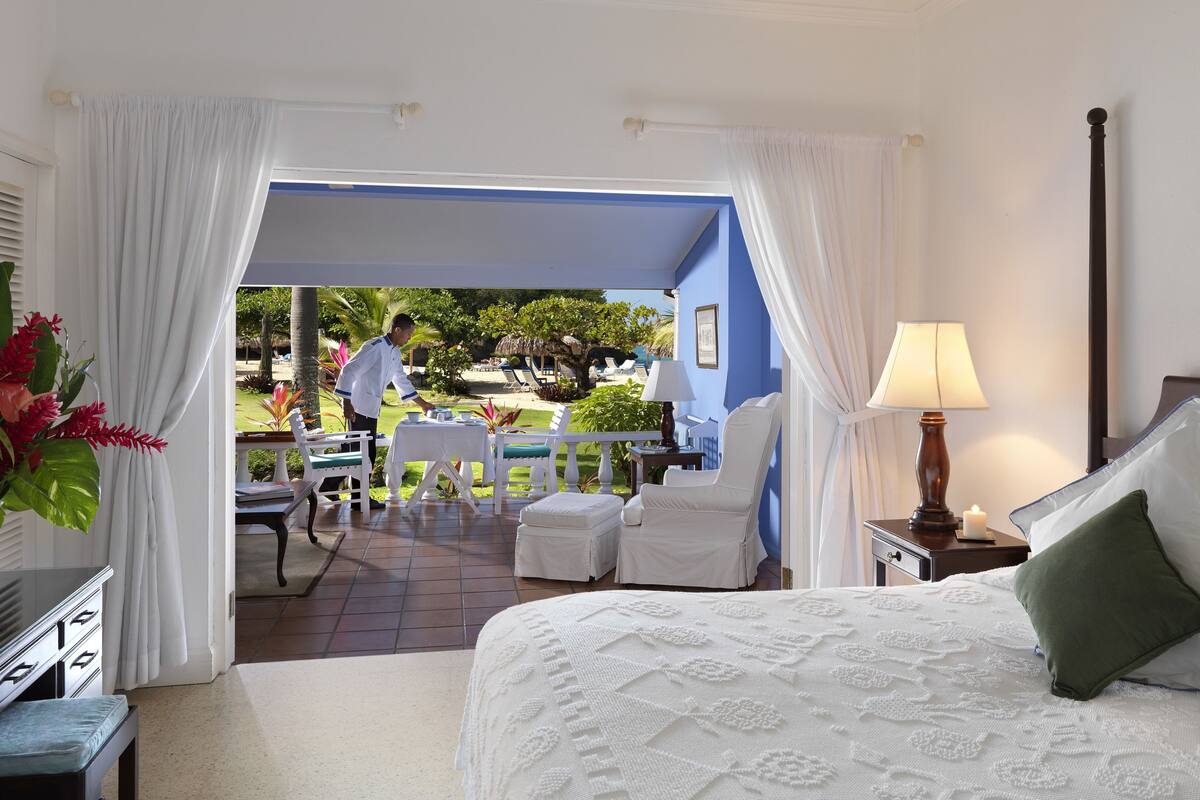 A hotel room with a bed, a nightstand, a lamp, and a view of the outdoor area with a table and chairs.