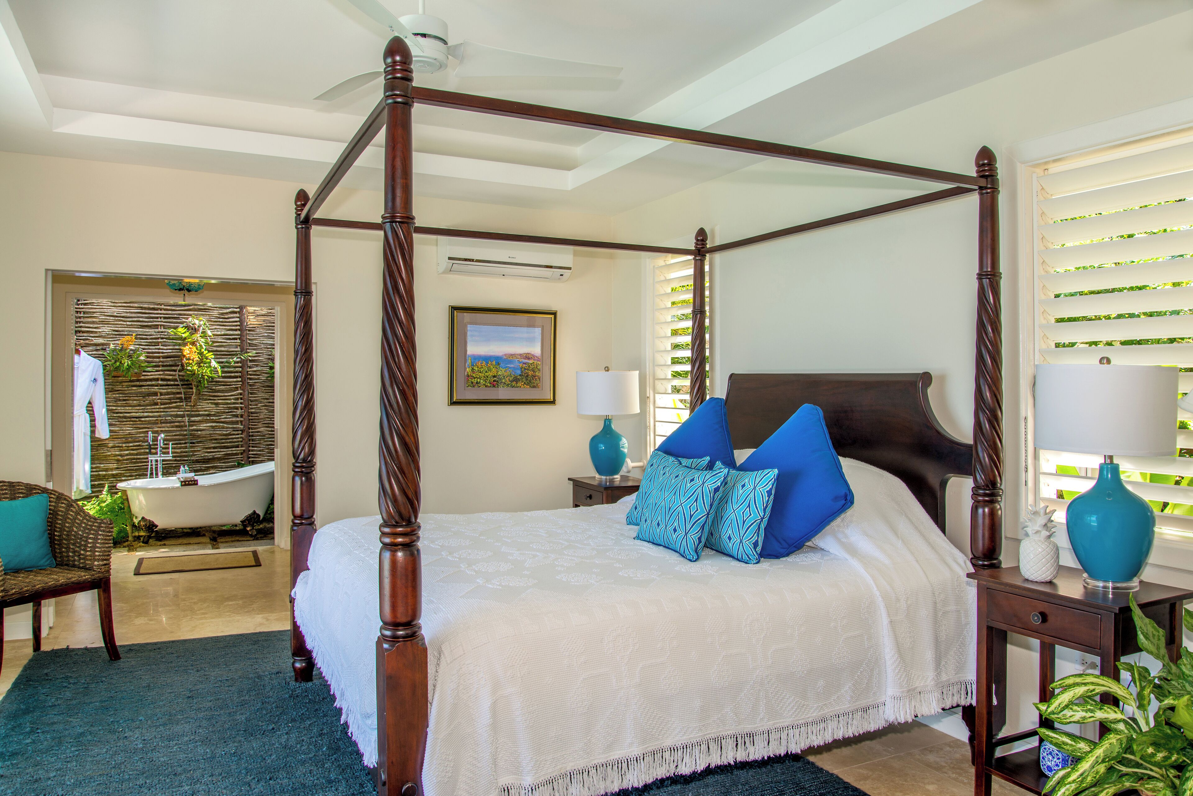 A four-poster bed with a white bedspread and blue pillows, a wooden nightstand with a lamp, and a view of a bathroom with a bathtub and plants.