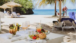 A beachside dining table with a salad, burger, and drinks, surrounded by lounge chairs and a palm tree.