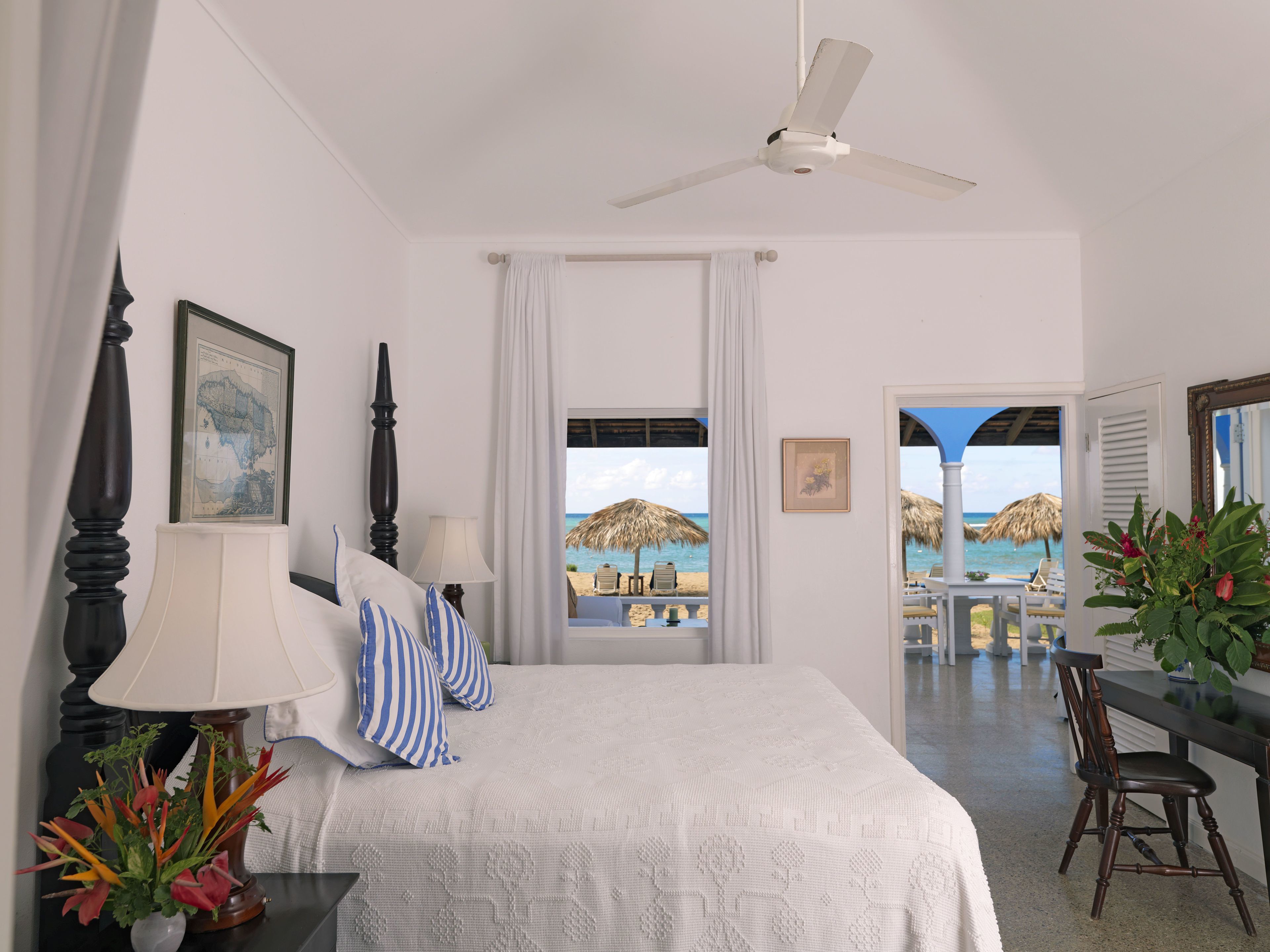 Habitación de hotel con cama, ventilador de techo, vista a la playa, zona de comedor y una planta en maceta.