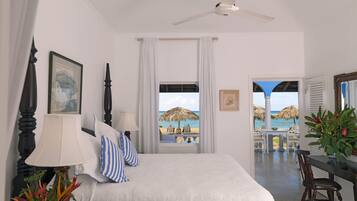 Habitación de hotel con cama, ventilador de techo, vista a la playa, zona de comedor y una planta en maceta.