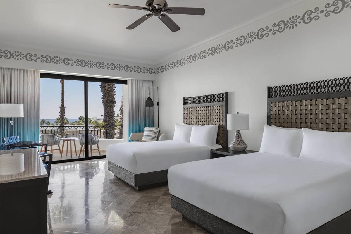 A hotel room with two beds, a desk, a chair, a balcony with a view, and a ceiling fan.