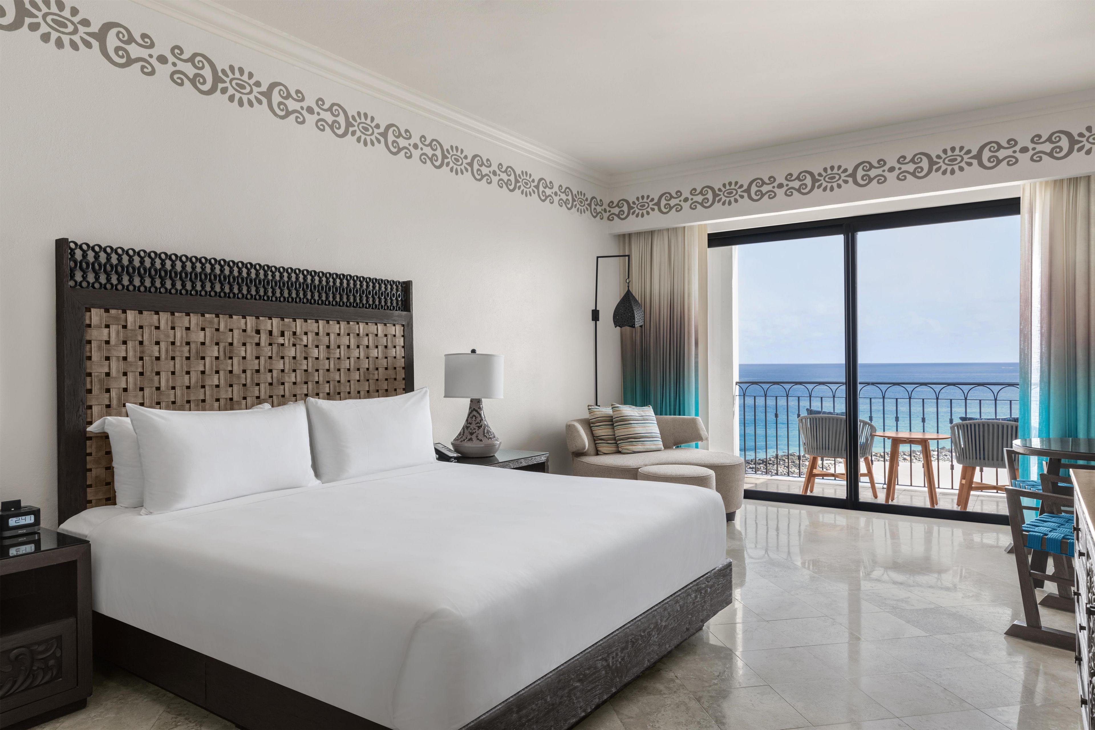A hotel room with a large bed, a nightstand, a lamp, a sofa, and a view of the ocean through a balcony.