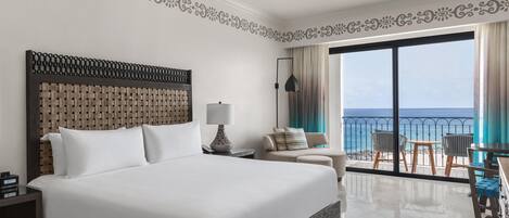 A hotel room with a large bed, a nightstand, a lamp, a sofa, and a view of the ocean through a balcony.