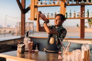 A bartender at a bar with a wooden interior, a view of the beach, and various bar tools and bottles on the counter. - Hilton Los Cabos (San José del Cabo)