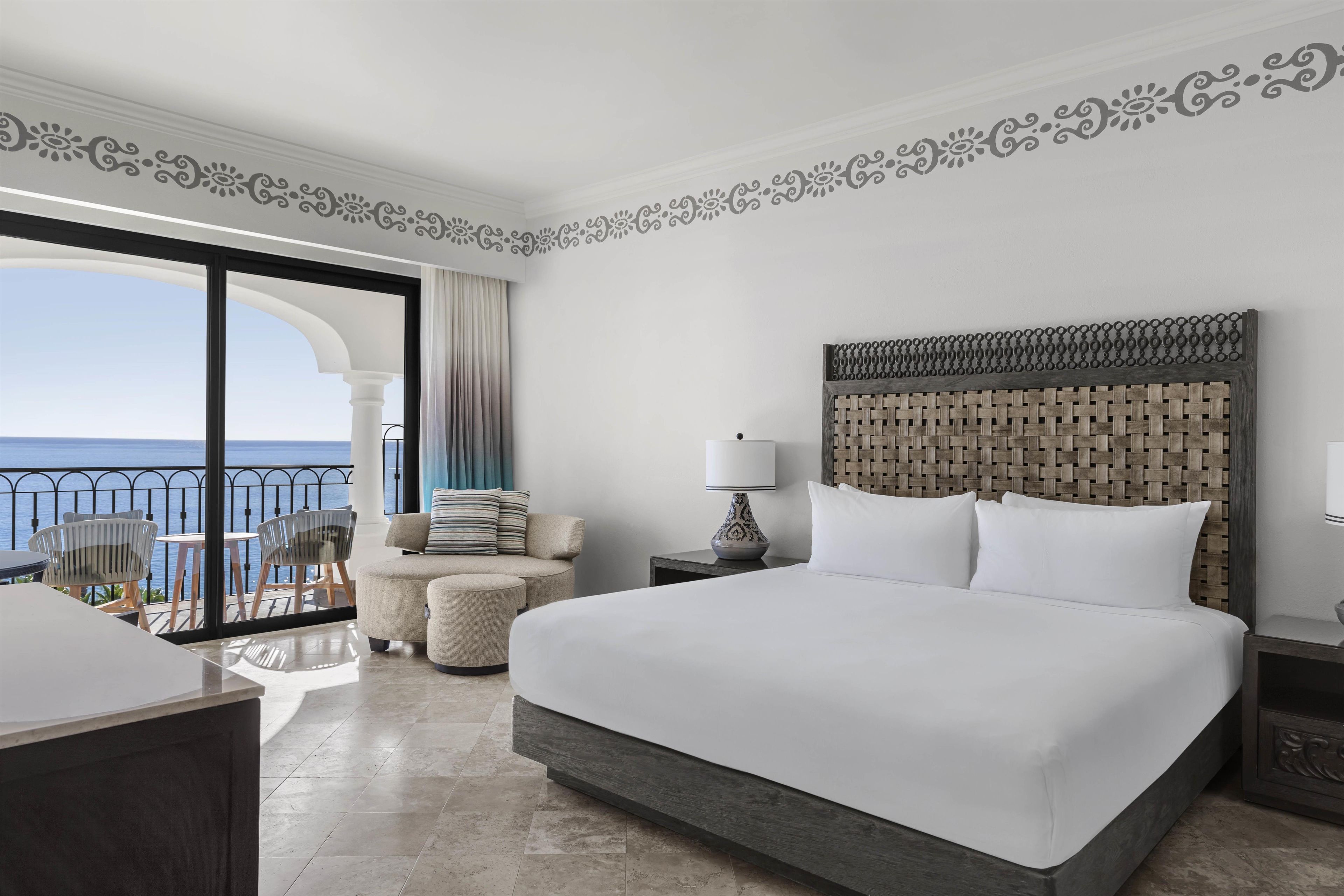 A hotel room with a large bed, a nightstand, a lamp, a chair, and a view of the sea through a balcony.