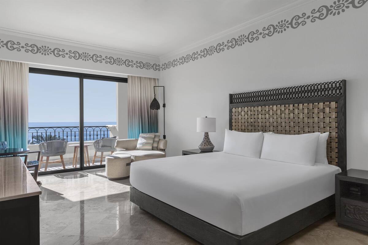 A hotel room with a large bed, a desk, a chair, a lamp, and a view of the sea.