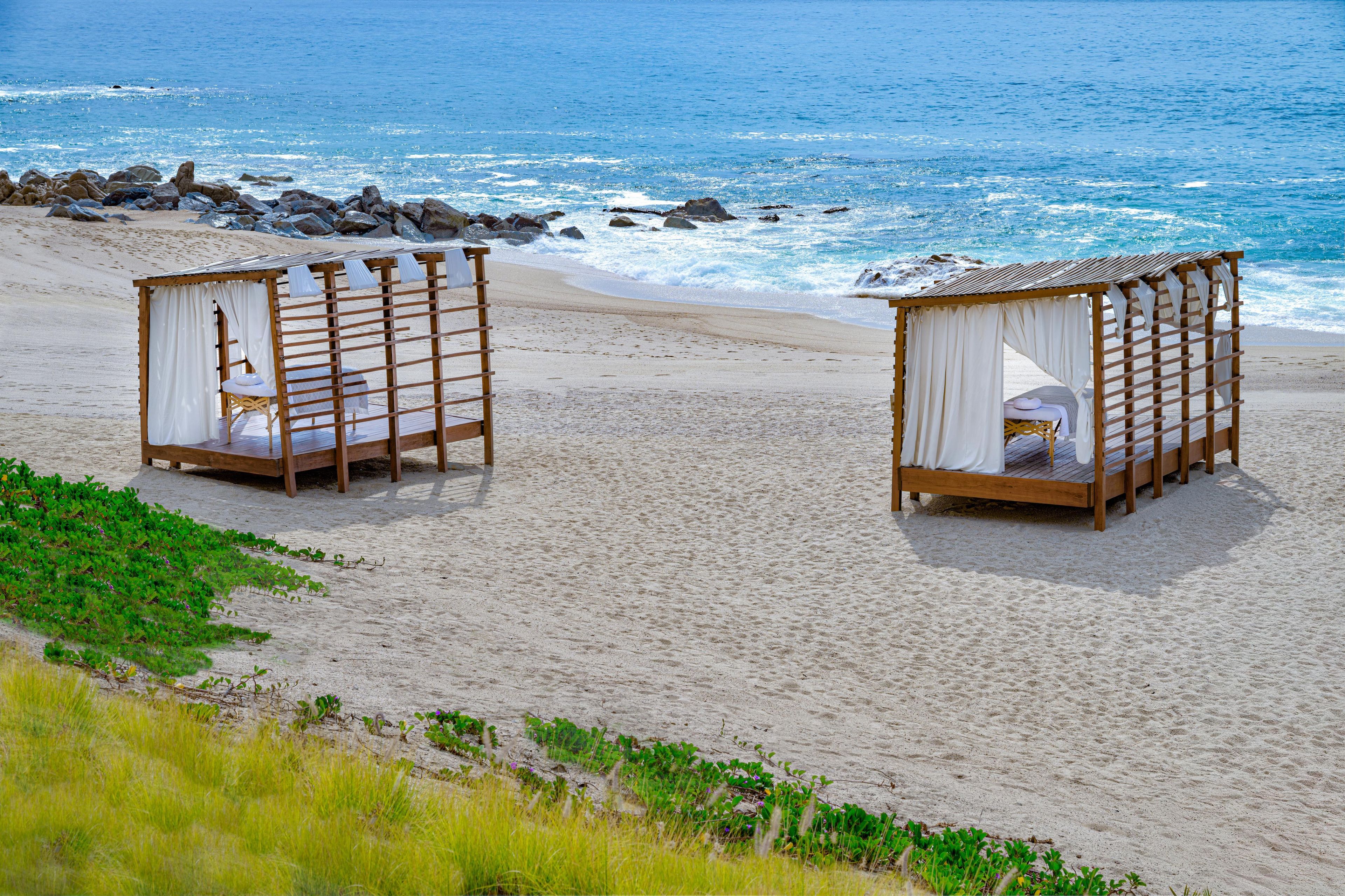 Two wooden bed structures with white curtains on a sandy beach with ocean waves in the background.