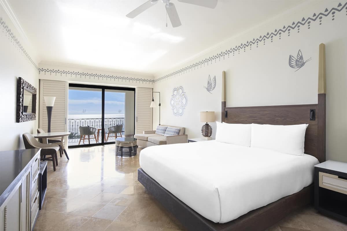 A hotel room with a large bed, a desk, a chair, a sofa, a balcony, and decorative wall art.