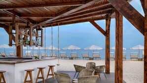 A wooden deck with bar stools, wicker chairs, and umbrellas overlooking a beach. - Hilton Los Cabos (San José del Cabo)