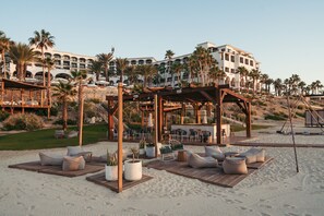 A beachside resort with outdoor seating, palm trees, and a large building in the background. - Hilton Los Cabos (San José del Cabo)