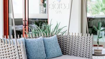Un sofá de mimbre con dos cojines azules, una pared con el texto "eforea SPA" y una ventana que da a zonas verdes.