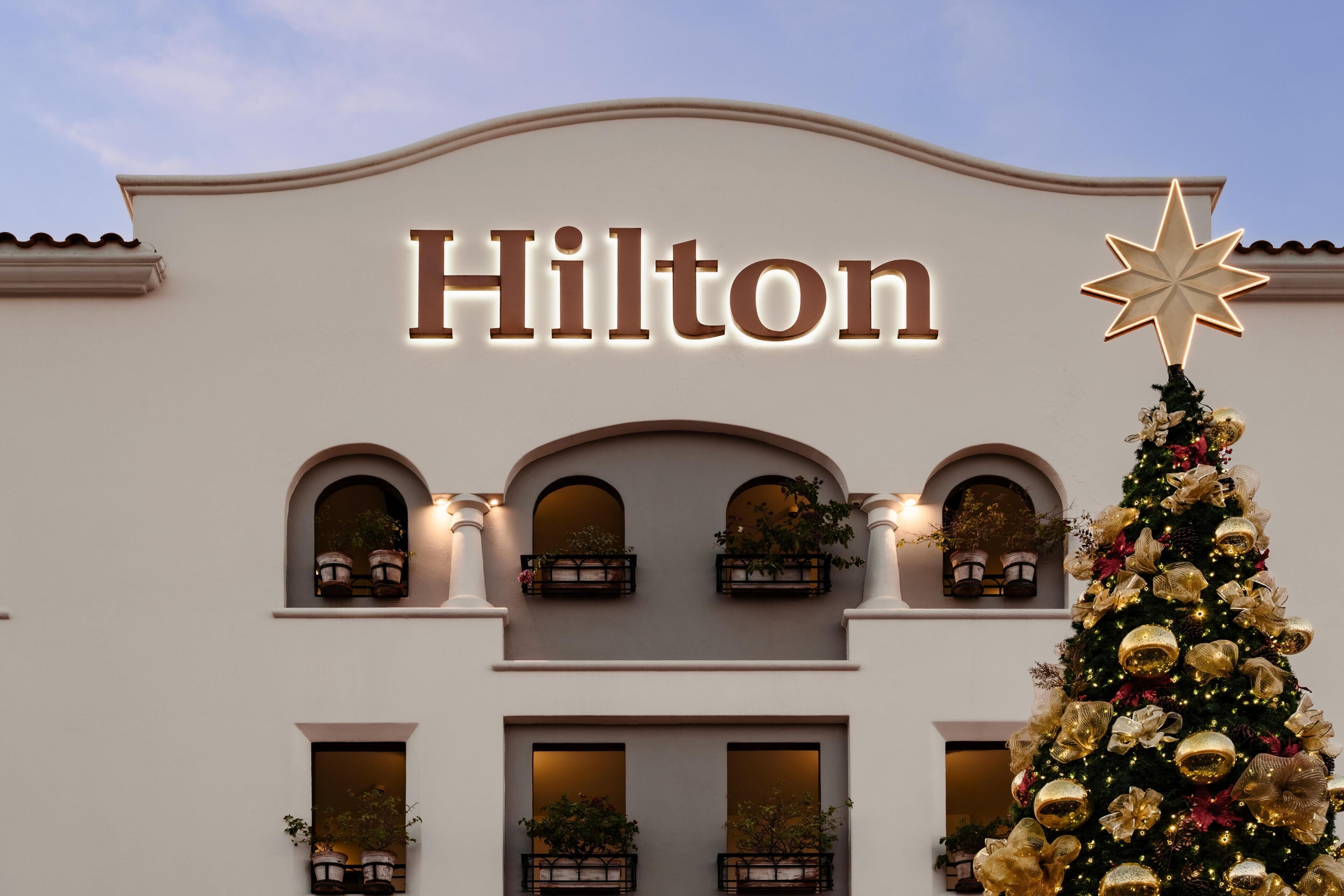 A building with the name 'Hilton' illuminated on the facade, balconies with potted plants, and a Christmas tree with ornaments.