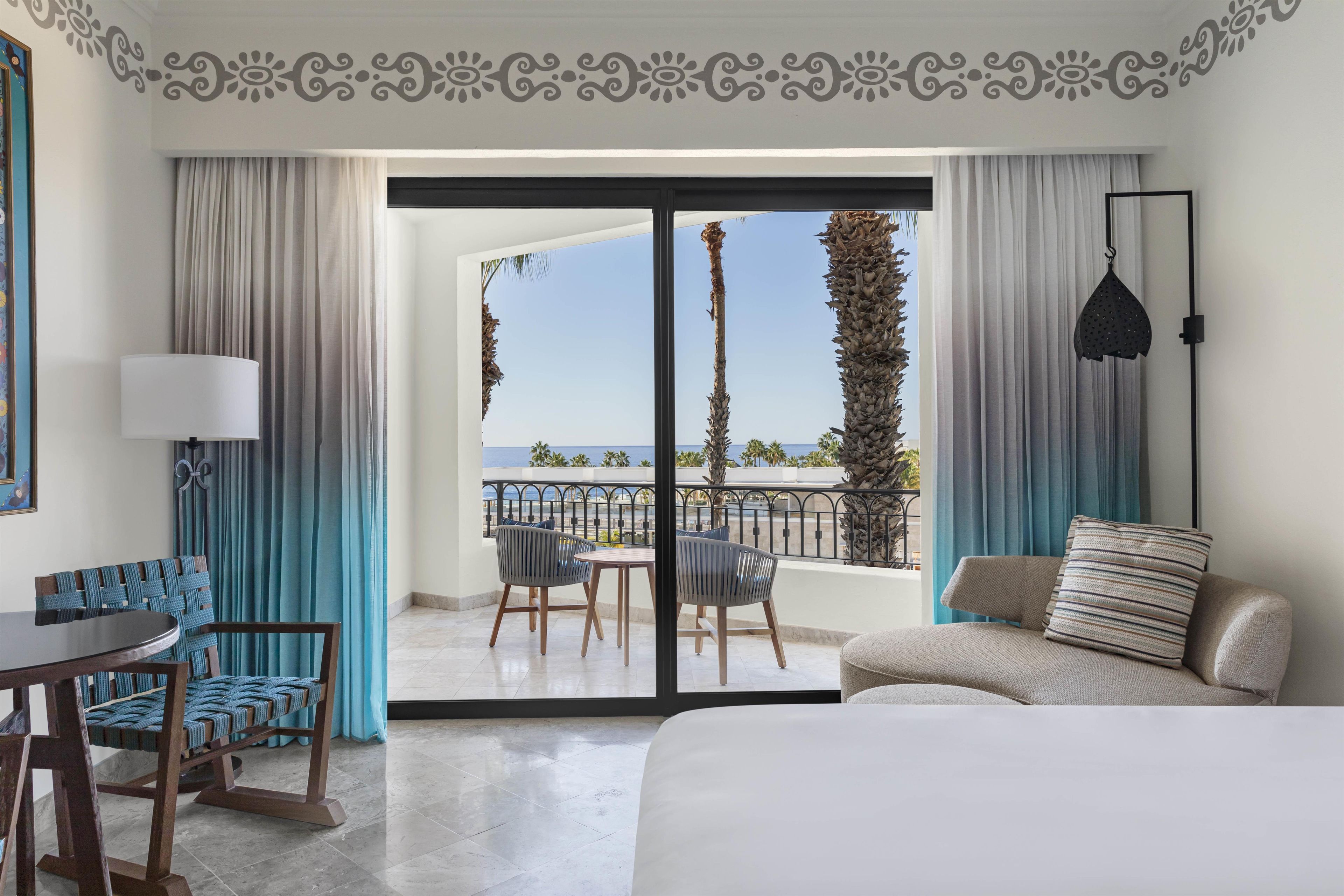A hotel room with a bed, a table, chairs, a balcony with a view of the sea, and a decorative ceiling design.