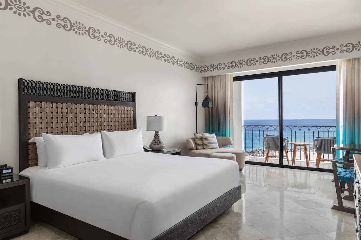 A hotel room with a large bed, a nightstand, a lamp, a chair, a table, and a balcony with a view of the ocean.