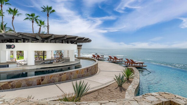 A poolside area with lounge chairs, a stone pool, palm trees, and a view of the ocean. - Hilton Los Cabos (San José del Cabo)