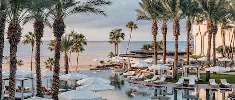 A resort pool area with lounge chairs and umbrellas, surrounded by palm trees and overlooking a beach.