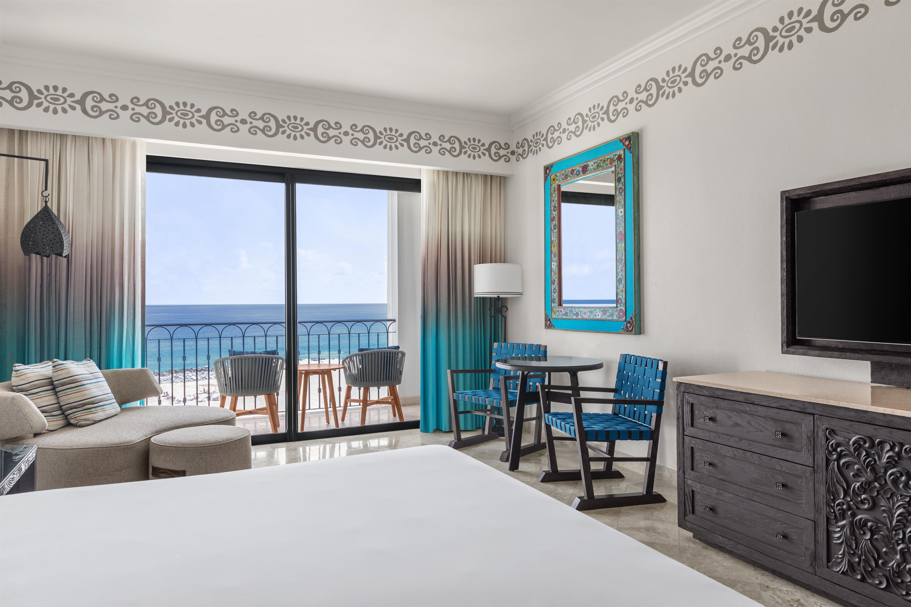 Habitación de hotel con balcón, cama, televisión, zona de comedor con sillas y un espejo decorativo.