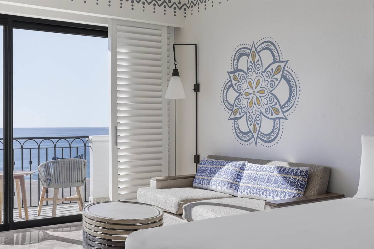 A hotel room with a balcony, a bed, a sofa, a patterned wall decoration, and a lamp.