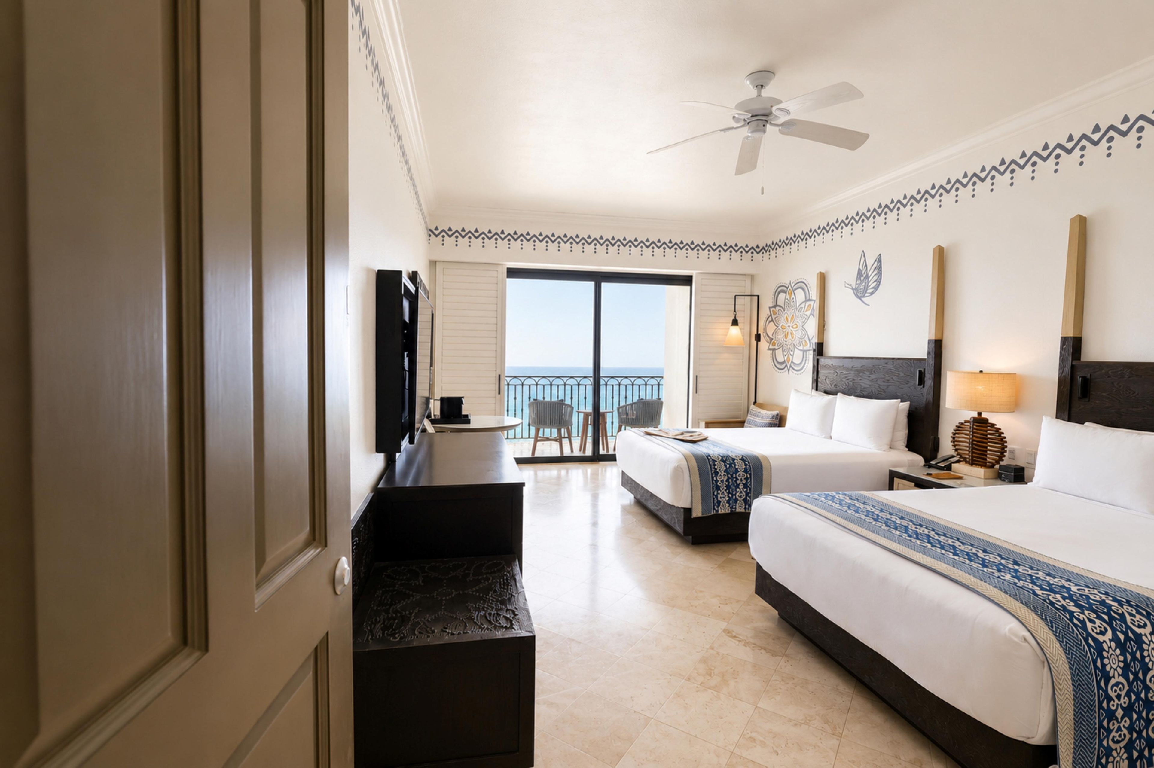 A hotel room with a bed, bedside tables, a desk, a chair, a balcony with a view, and a ceiling fan.