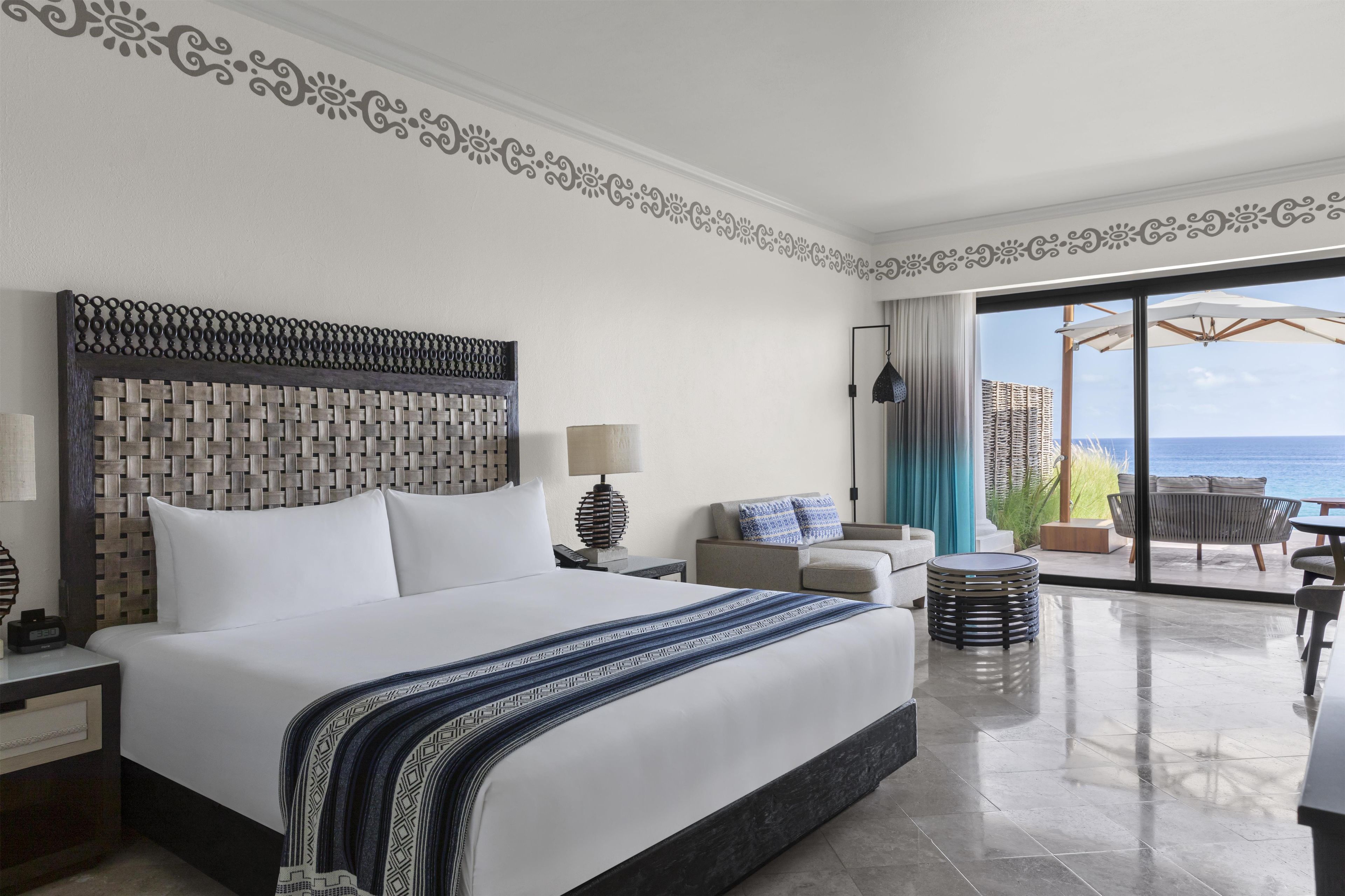 Ein Hotelzimmer mit einem großen Bett, Meerblick, einem Balkon mit Sitzgelegenheiten und einem dekorativen Wandmuster.