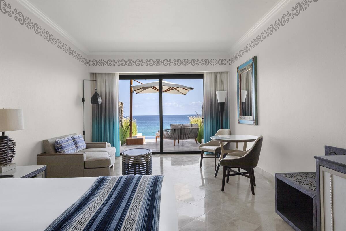 A hotel room with a bed, a sofa, a dining table, a chair, a lamp, and a view of the ocean through a glass door.