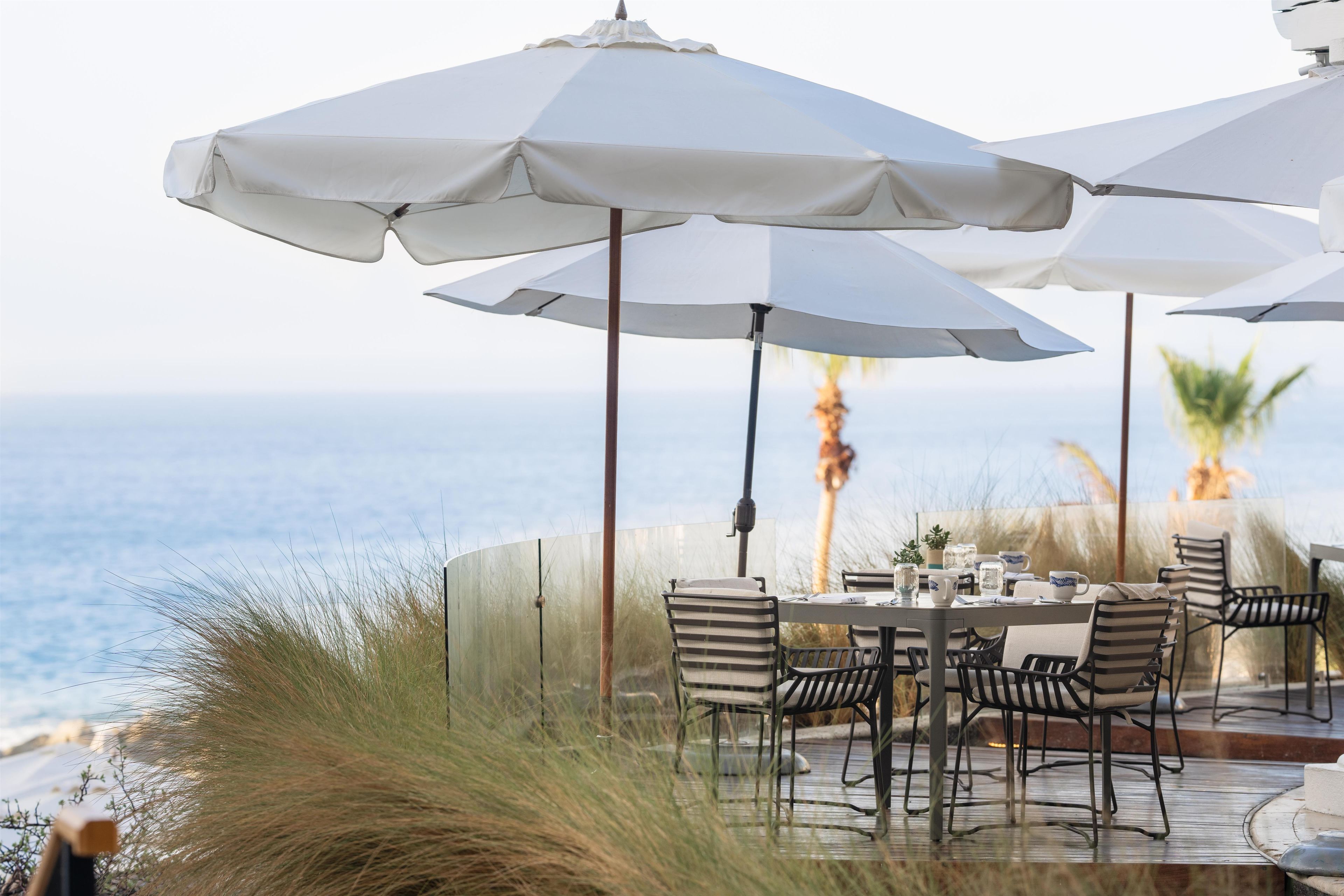 Un área de comedor al aire libre con mesas y sillas, sombrillas blancas y vista al océano.