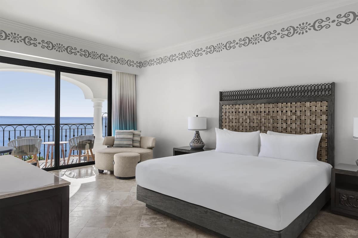 A hotel room with a large bed, a balcony with a view of the sea, a small round table, and a sofa.