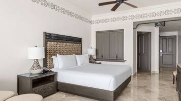 A hotel room with a large bed, two bedside tables with lamps, a ceiling fan, a wardrobe, and a door leading to another room.