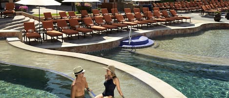 A pool area with lounge chairs and umbrellas, a clear pool, and a building with balconies in the background.
