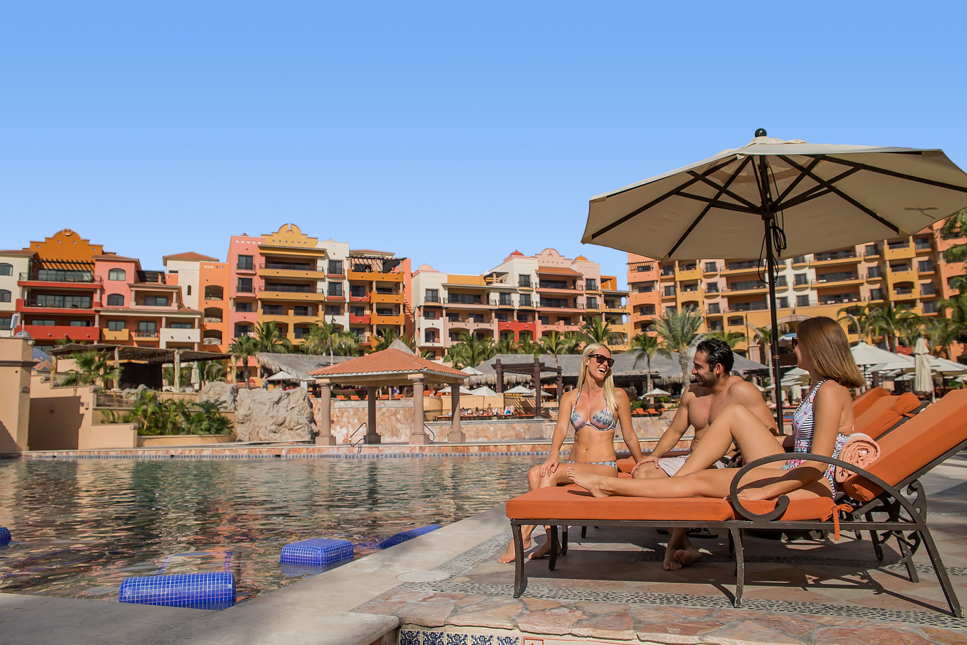 Zona de piscina con sillas de descanso y sombrillas, rodeada de edificios de varios pisos con balcones.