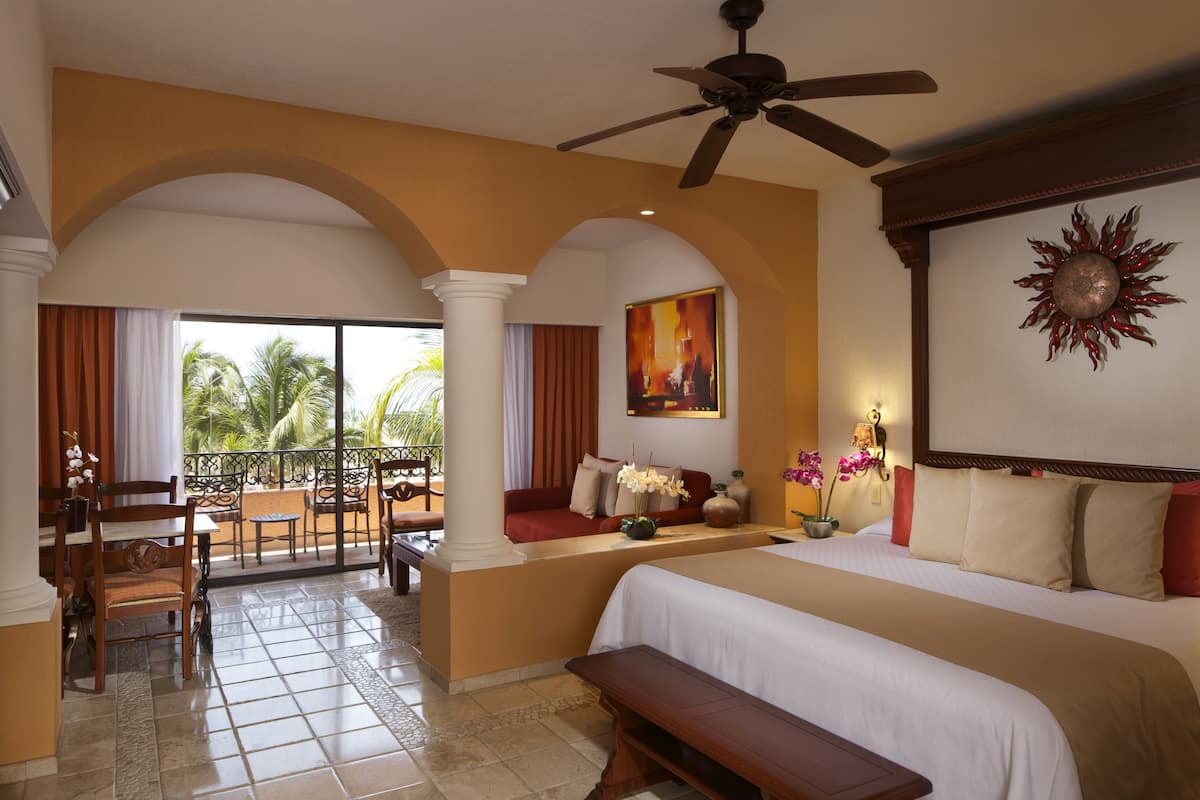 Une chambre d’hôtel avec un grand lit, un ventilateur de plafond, un balcon avec des sièges et une vue sur les palmiers.