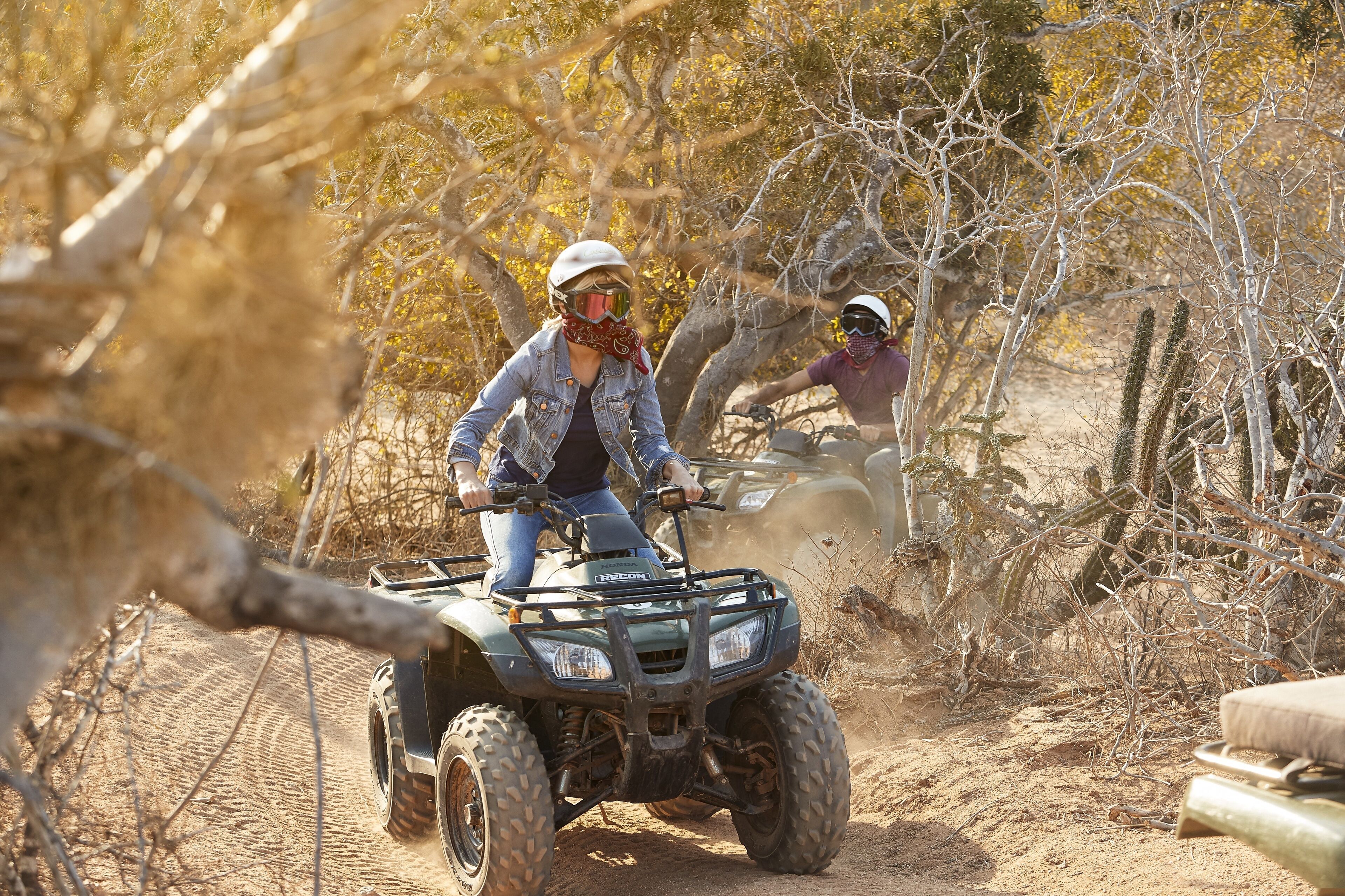 Two people riding an ATV on a dirt path surrounded by dry vegetation and trees.