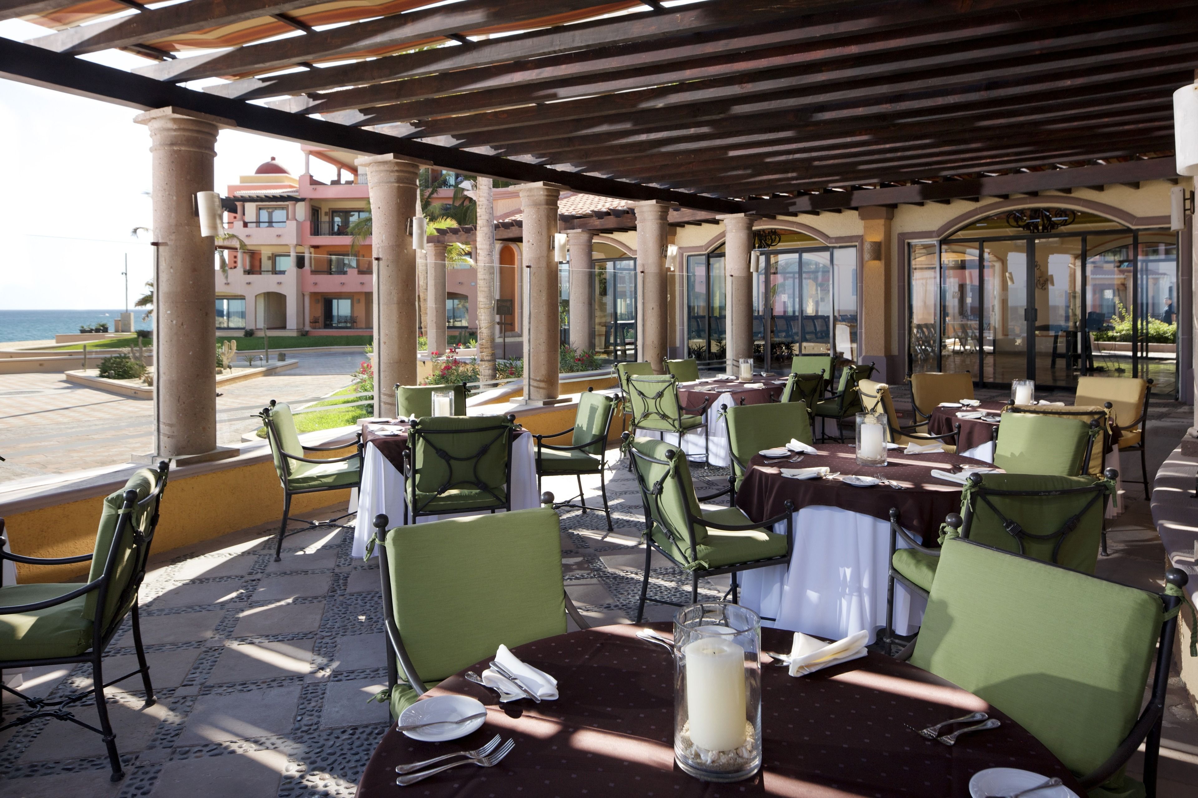 An outdoor dining area with tables and chairs, a view of the ocean, and a building with a pink facade in the background.