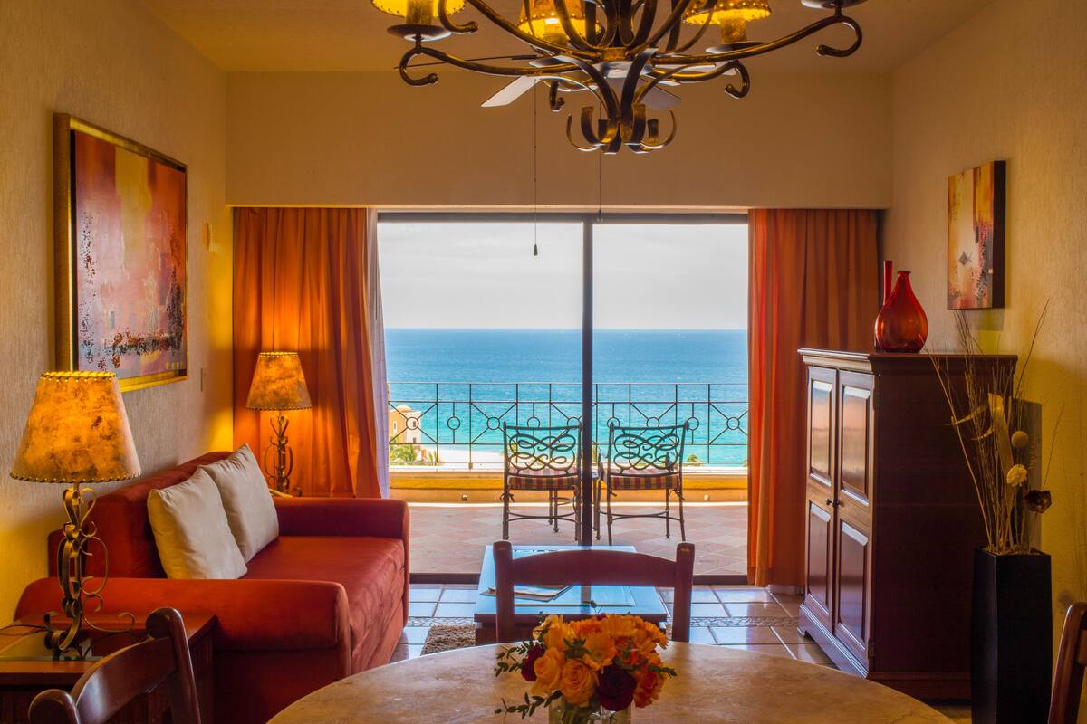 Une chambre d’hôtel avec un balcon, une table à manger décorée de fleurs, un canapé rouge et un lustre.