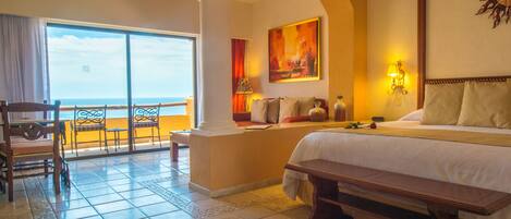 A hotel room with a large bed, a bench, a desk, and a view of the ocean.