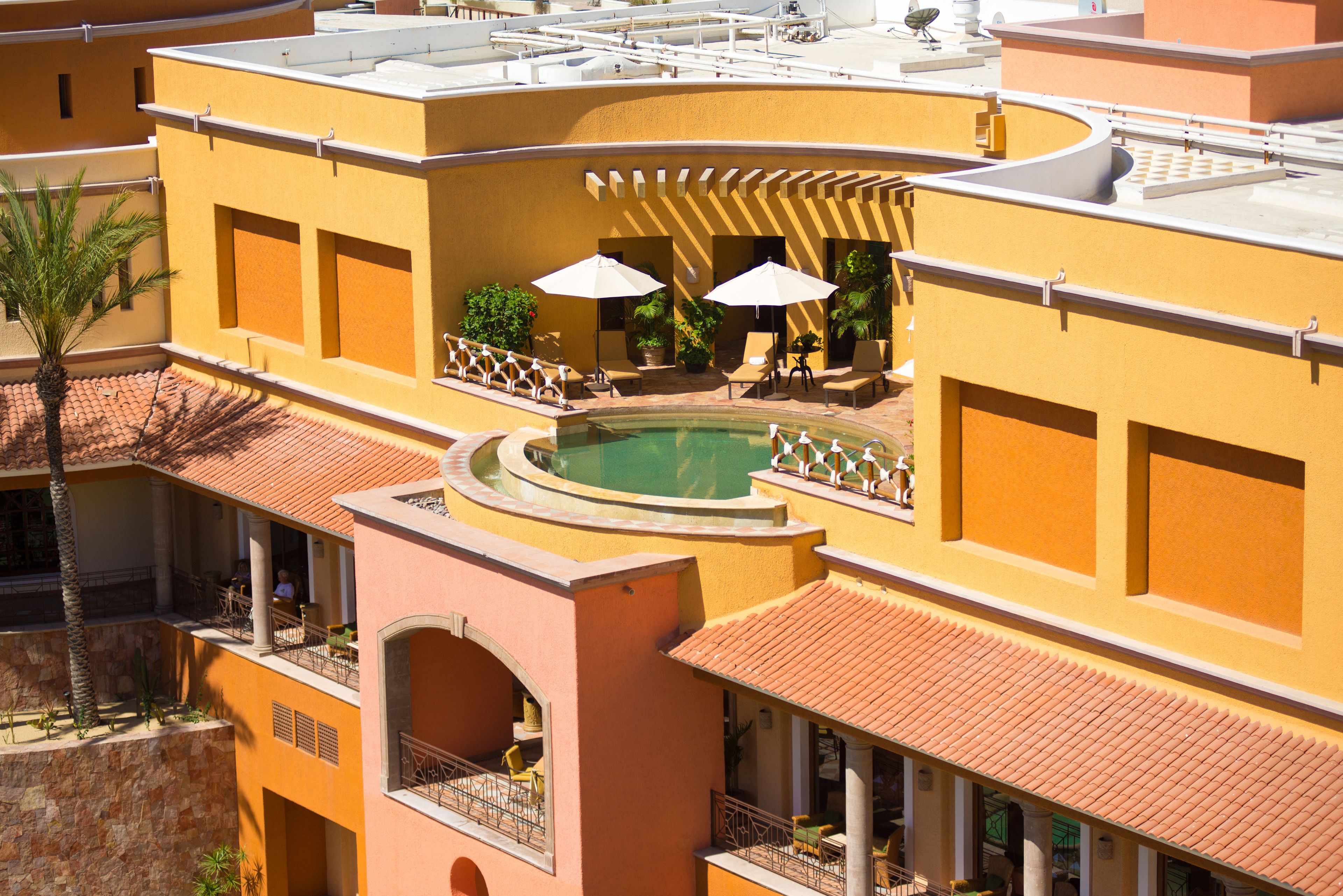 A multi-level hotel with a rooftop pool, outdoor seating with umbrellas, and a palm tree.