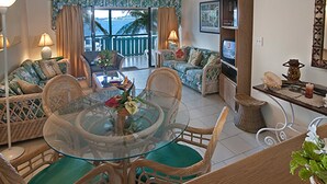 A living room with a glass-top dining table, wicker furniture, and a view of the ocean through large windows.