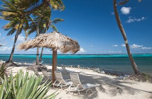 Una playa con una sombrilla de techo de paja, palmeras y sillones blancos para tomar el sol.