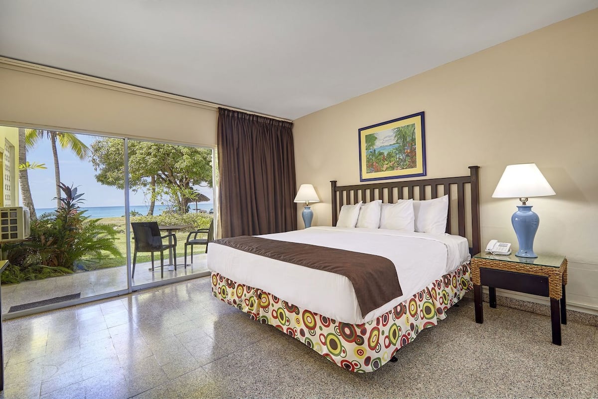 A hotel room with a bed, two bedside lamps, a painting on the wall, and a view of the ocean through large windows.