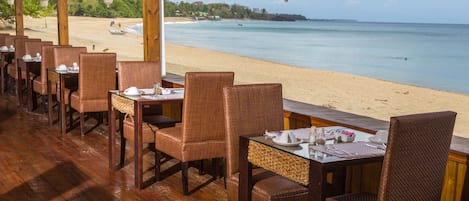 An outdoor dining area with wicker chairs and tables, overlooking a beach and ocean.