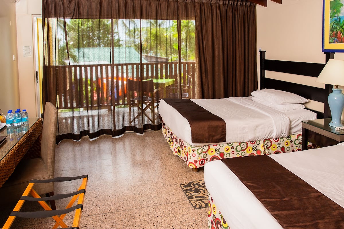 A hotel room with two beds, a desk with a chair, a table with chairs outside, and a view of the outdoors.