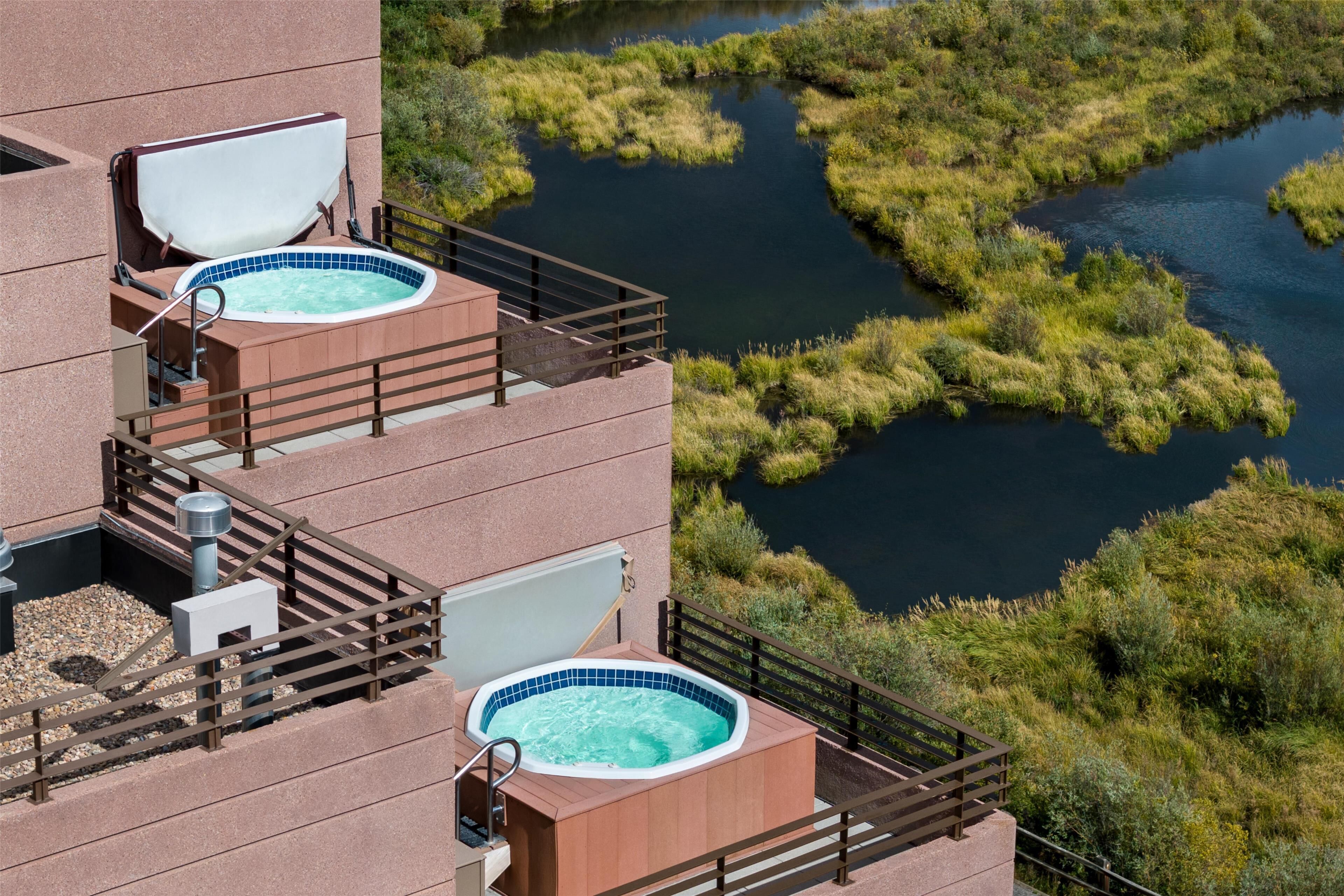 A hotel with two outdoor hot tubs on a balcony, surrounded by a natural landscape with a river and greenery.