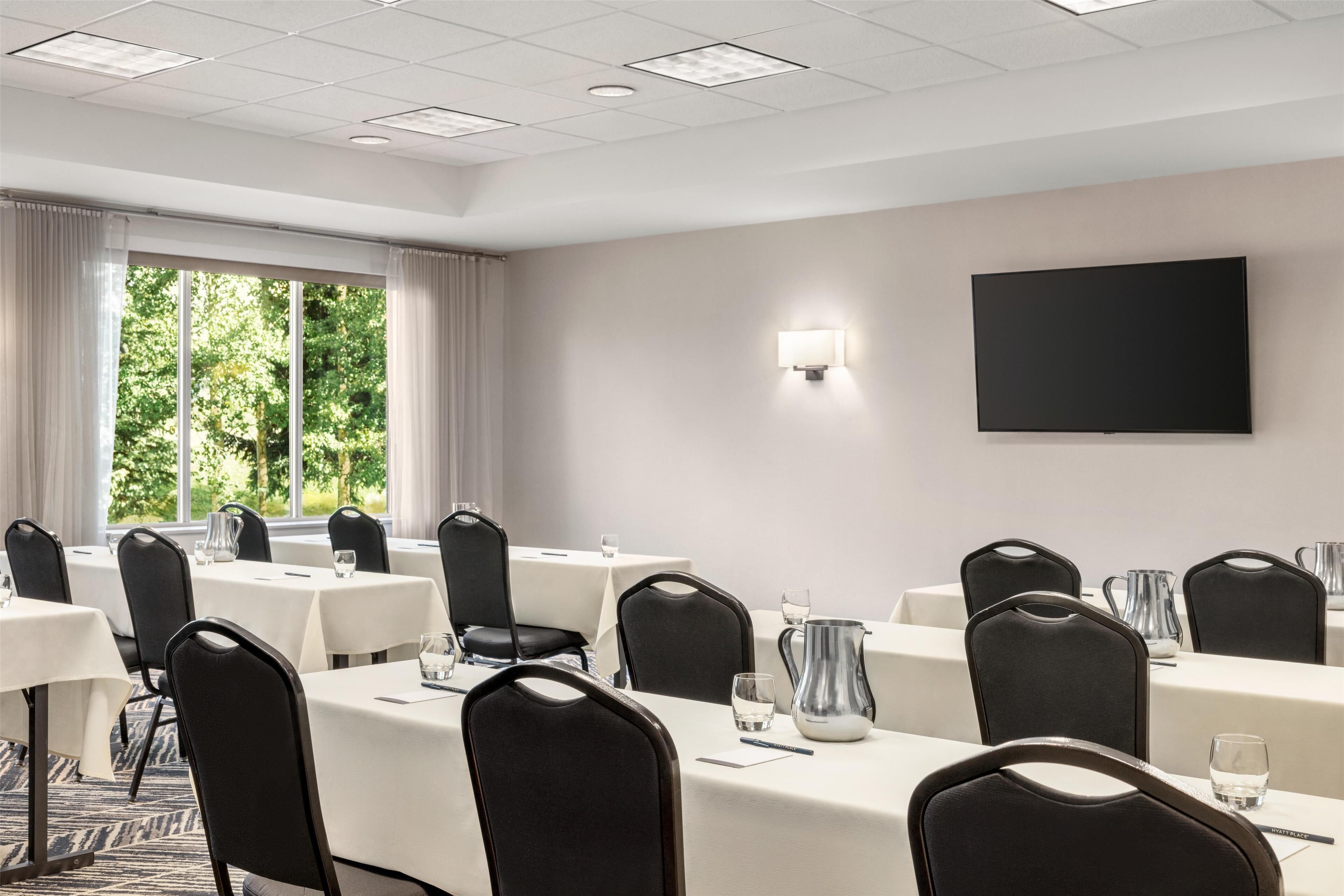 A conference room with tables, chairs, a TV, and a window with curtains.