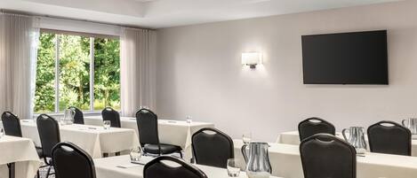 Un salón de conferencias con mesas, sillas, una televisión y un ventanal con cortinas.