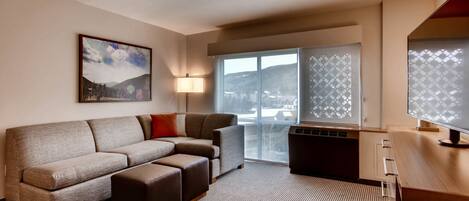 Une chambre d’hôtel moderne avec un canapé gris, un petit pouf, un bureau avec un ordinateur, une lampe et une grande fenêtre donnant sur les montagnes.