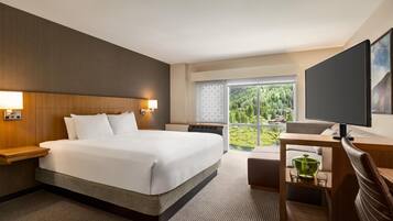 A hotel room with a large bed, a desk with a computer, a TV, and a view of a forest through the window.