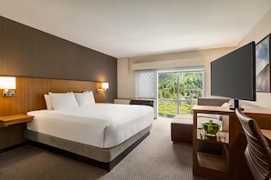 A hotel room with a large bed, a desk with a computer, a TV, and a view of a forest through the window.