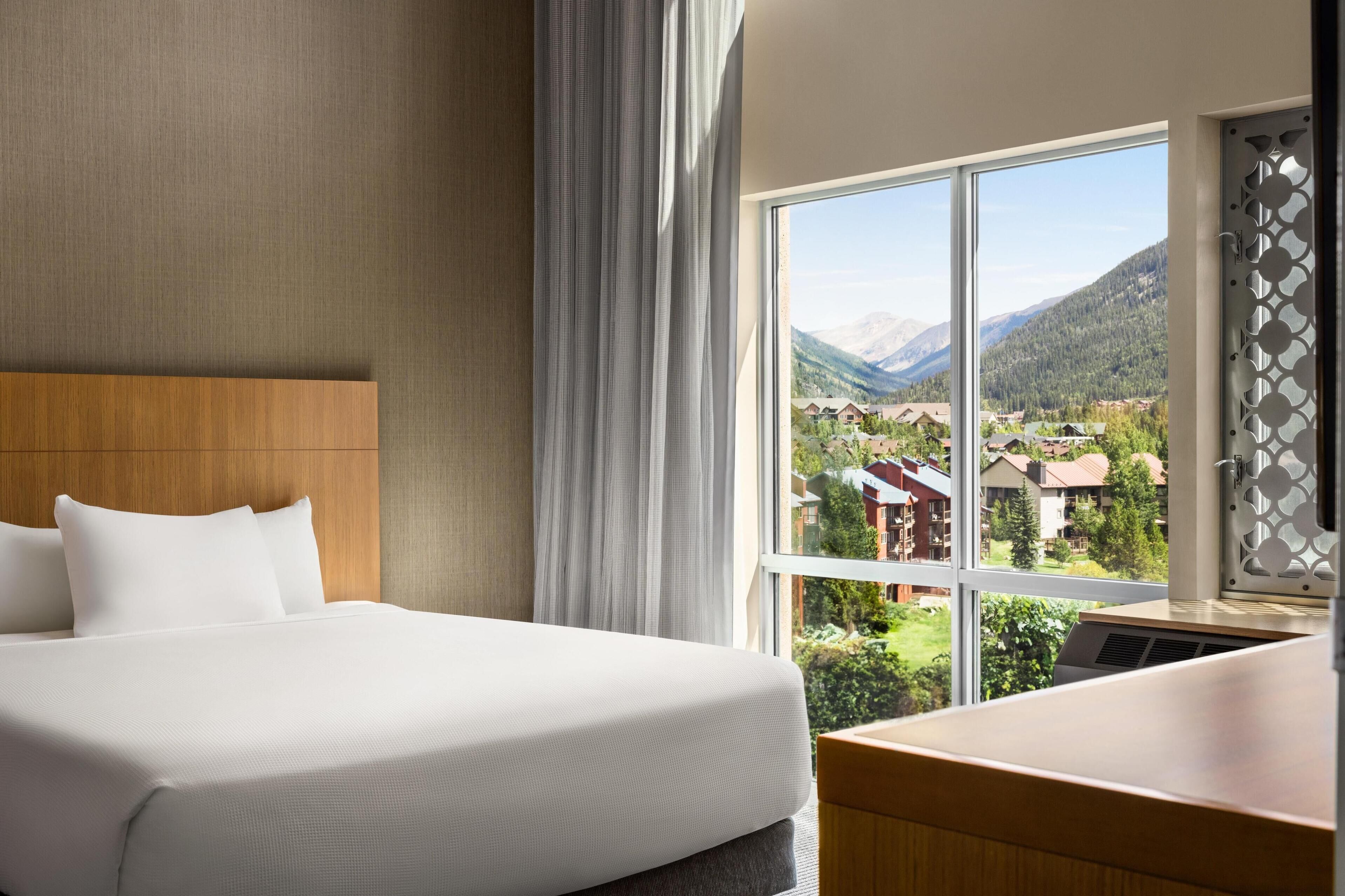 A hotel room with a large bed, a wooden headboard, a view of mountains and houses through a large window, and a wooden desk.